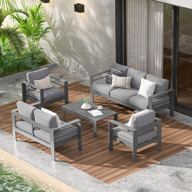 Outdoor patio furniture set on a wooden deck with plants and a building in the background.