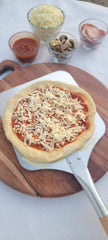 Uncooked pizza with cheese on a wooden board with ingredients in small bowls.
