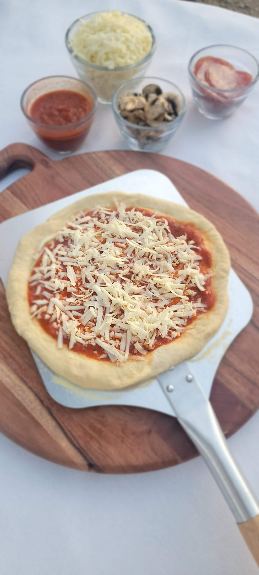 Uncooked pizza with cheese on a wooden board with ingredients in small bowls.