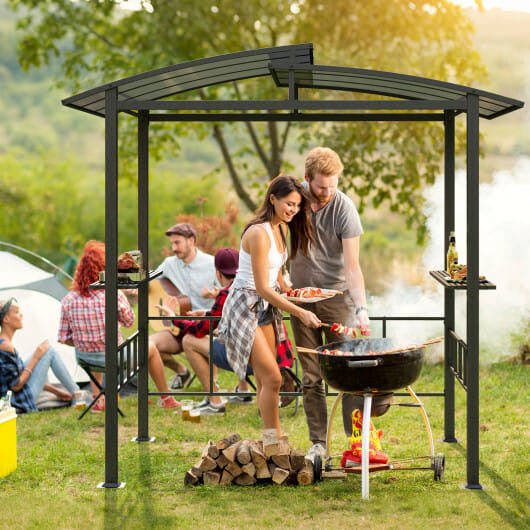 People grilling outdoors under a metal gazebo with a scenic background
