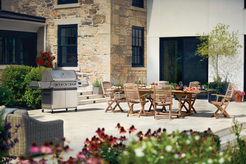 Outdoor patio with wooden furniture, grill, and flowers in front of a stone building.