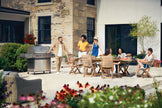 Family and friends gathered around a grill and dining table in a backyard setting.