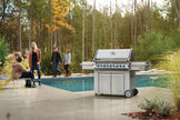 Stainless steel grill on a patio with people near a pool and trees in the background