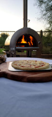 Wood-fired pizza being cooked in an outdoor oven with a scenic background.